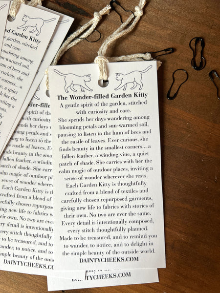 Product description card for 'The Wonder-filled Garden Kitty' on a wooden surface with twine and clips.