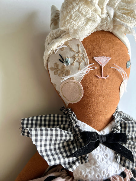 Textile art doll of a cat head with lace and checkered details on a plain background