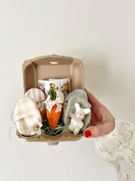 Hand holding a small cardboard egg carton with decorative Easter eggs and a bunny figure on a light background