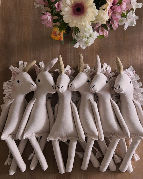 Row of white unicorn dolls with decorative elements on a wooden surface with flowers in the background.